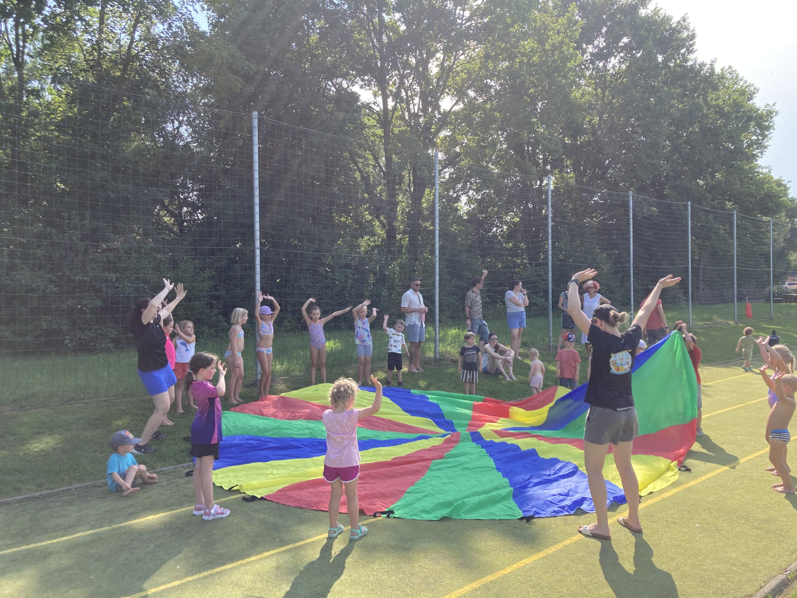 Kinder und Erwachsene spielen mit einem bunten Schwungtuch auf einem Sportplatz in Österreich.